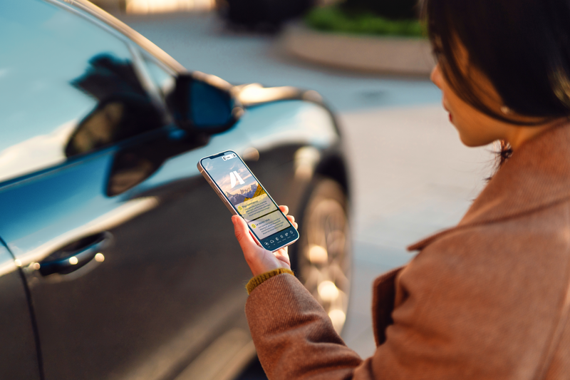 Over the shoulder view of young woman using smartphone against an electric car. Car sharing service. Transportation and technology concept.
