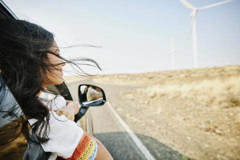 Girl with her head out the car window, hair blowing in the wind, passing wind turbines while driving on a road.